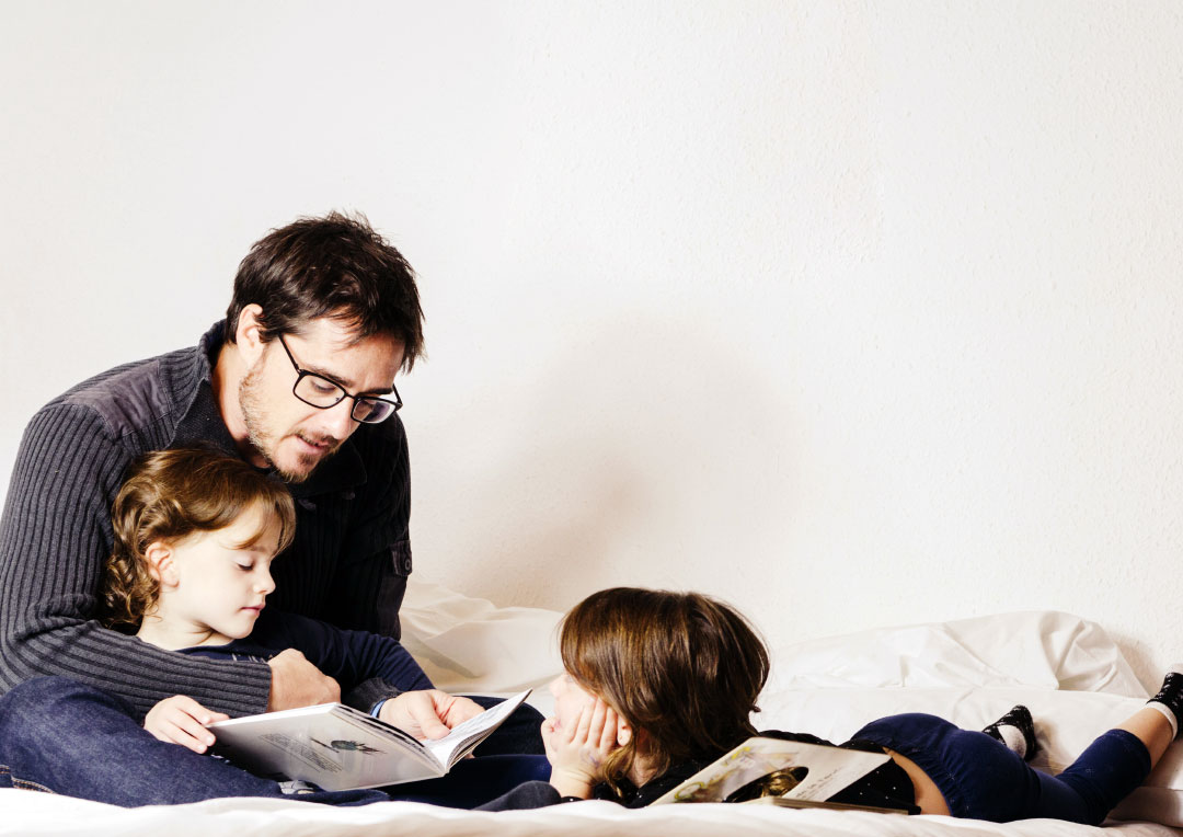 pai sentado na cama lendo pra dois filhos pequenos