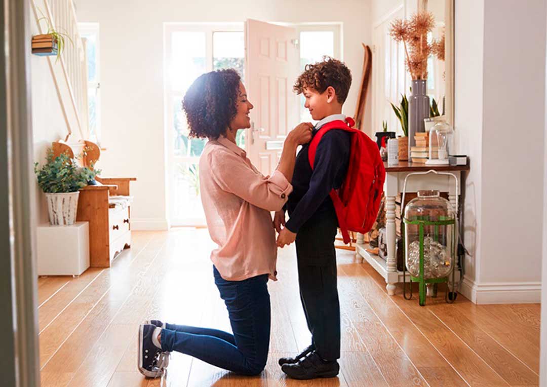 mãe de joelho arrumando a blusa do filho com mochila de escola