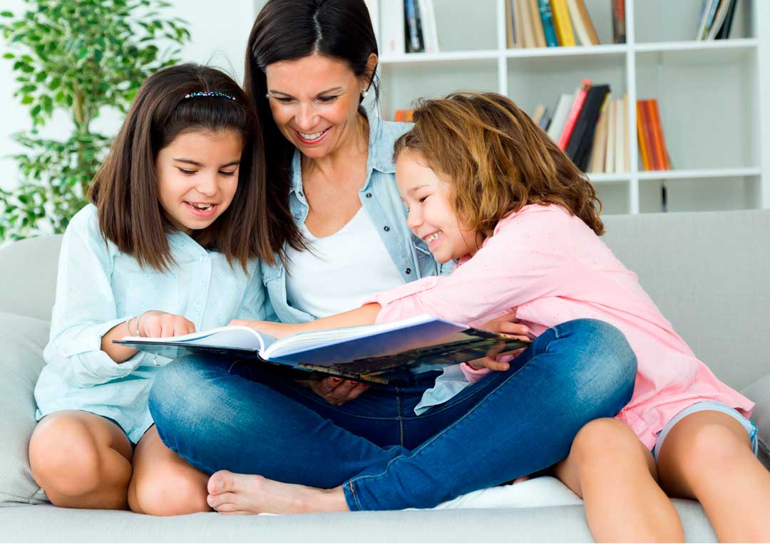 mãe com duas filhas felizes sentadas no sofá fazendo leitura