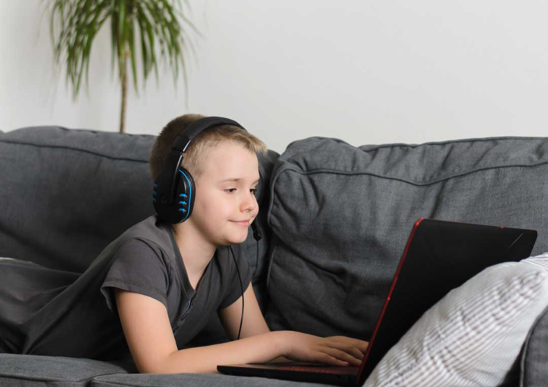 menino com fone de ouvido, deitado no sofá e usando um notebook